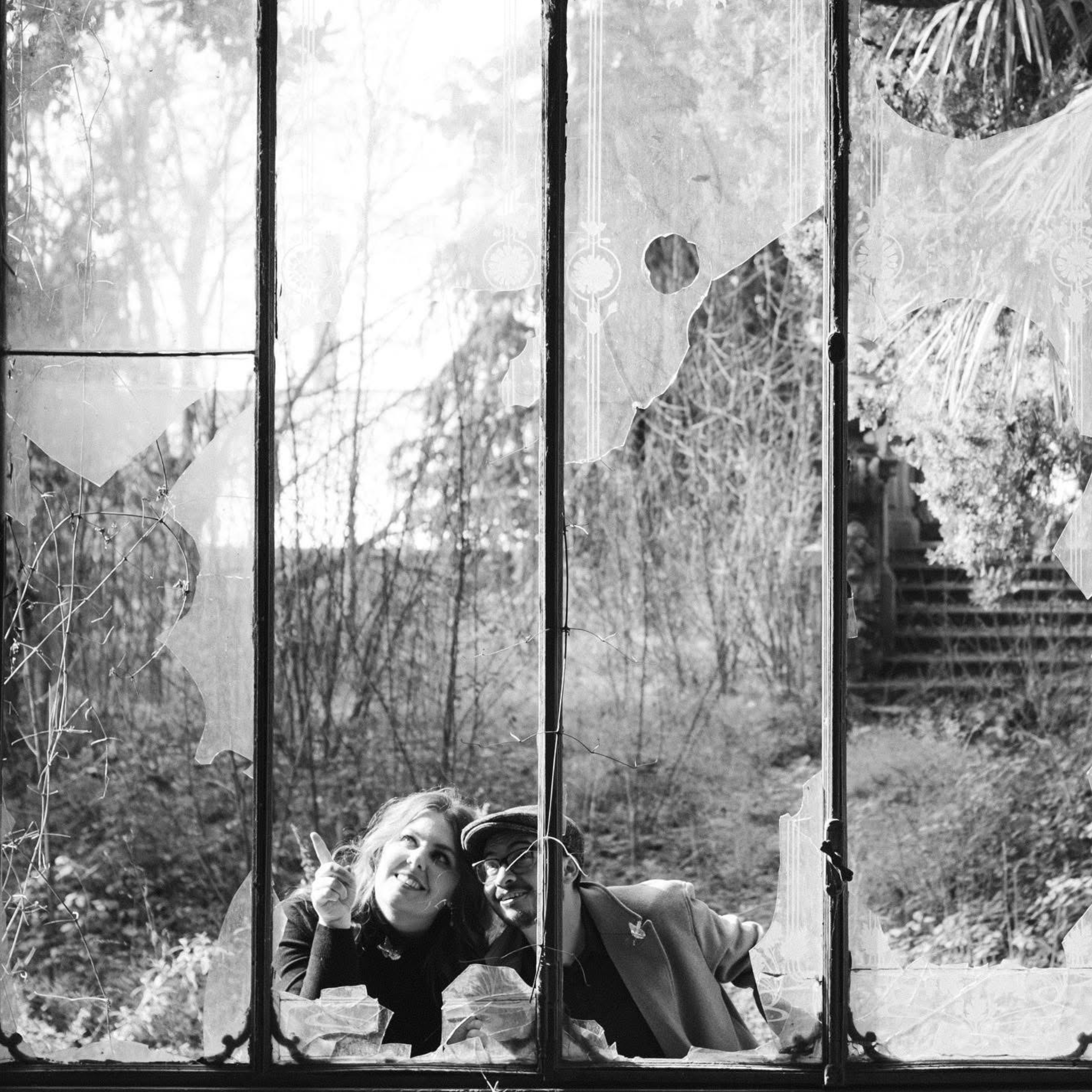 A black-and-white photo of a couple, smiling and leaning close together while looking up through a broken glass window. They are framed by the jagged edges of the shattered panes, surrounded by overgrown foliage and soft light. The setting appears to be an abandoned greenhouse or building in Italy, creating a poetic and intimate atmosphere.